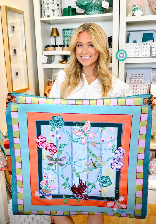 A smiling blonde woman in a white blouse holds up mahjong mat featuring a colorful butterfly and floral design by Jennifer Poe Art. The product has a turquoise border with pink, green, and yellow stripes, an orange inner border, and a light blue center with embroidered butterflies in pink, purple, green, and red.